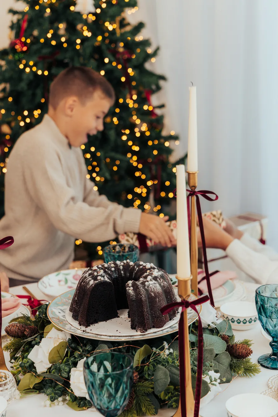 A boy reaching across a laid table, a chocolate bundt cake and tall candles in front of him