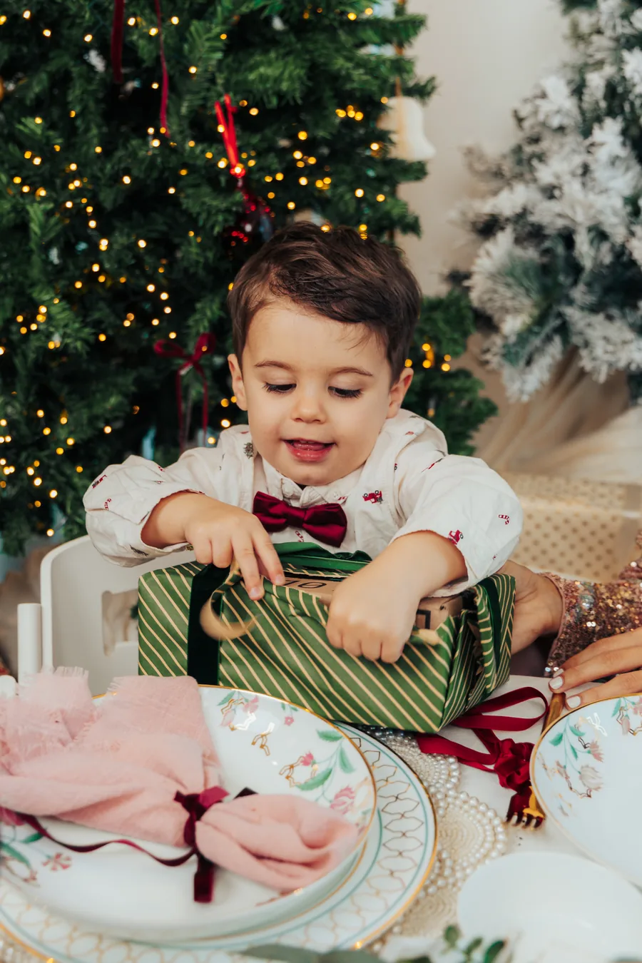 A small boy in a white shirt and bow tie tearing into a green wrapped gift at the table