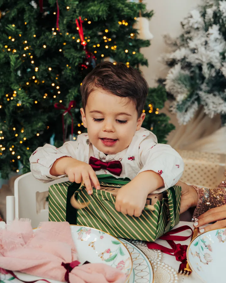 A small boy in a white shirt and bow tie tearing into a green wrapped gift at the table