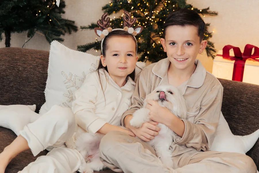 Two boys in cream pyjamas on the sofa holding a small white dog, the tree behind them