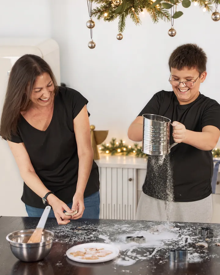A mother laughing as her son sifts flour over the table in the Christmas Kitchen