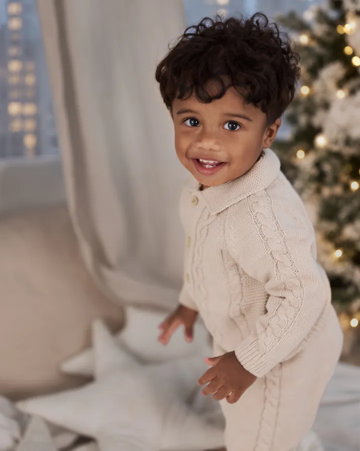 A smiling toddler in a cream knitted suit among the stars and snowy trees of Christmas Morning