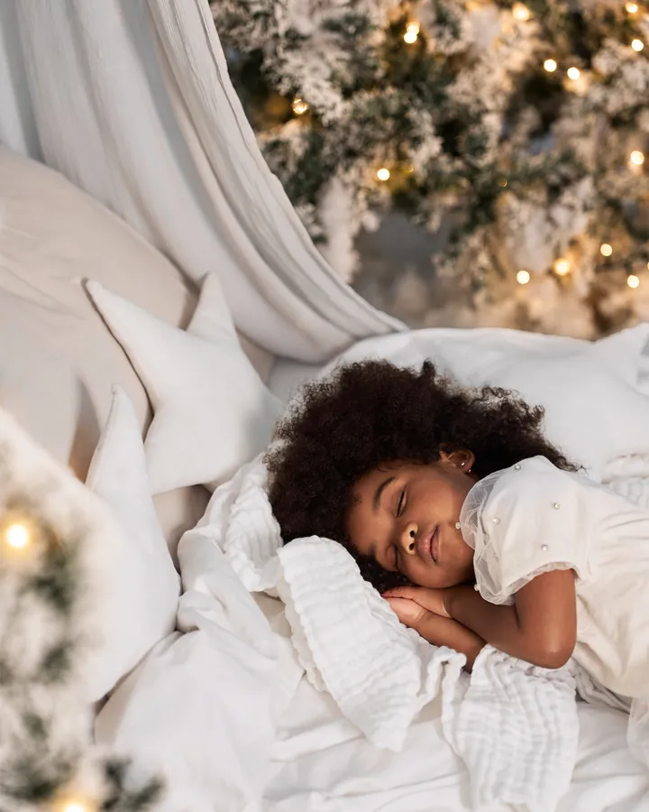 A little girl asleep among white pillows and soft stars, fairy lights behind her