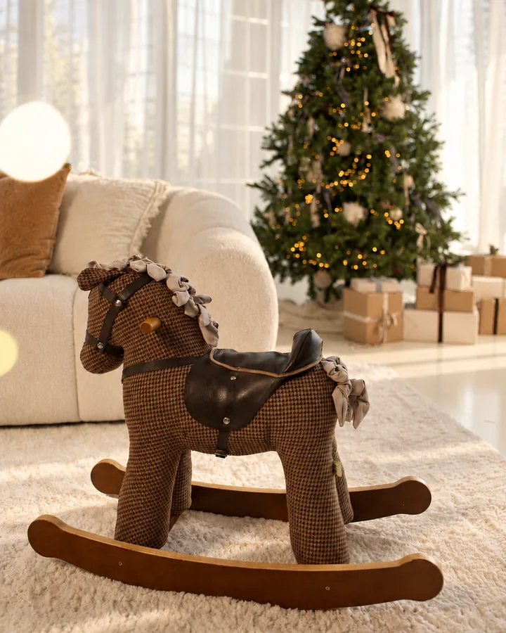 A rocking horse on a cream rug in the Christmas Room, a lit tree and wrapped presents behind it