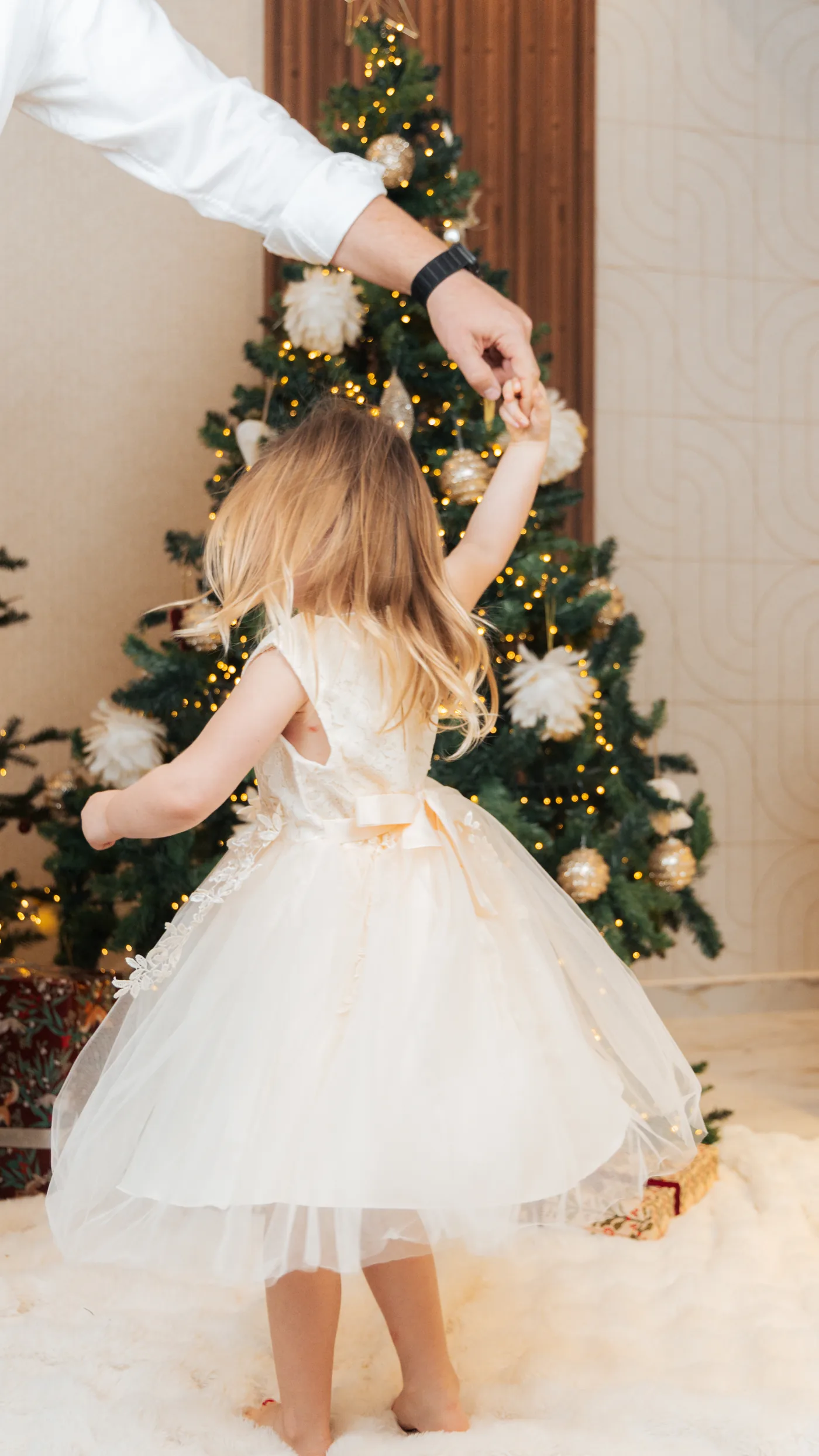 A small girl in a cream tulle dress spinning under her father&rsquo;s hand in front of a lit tree